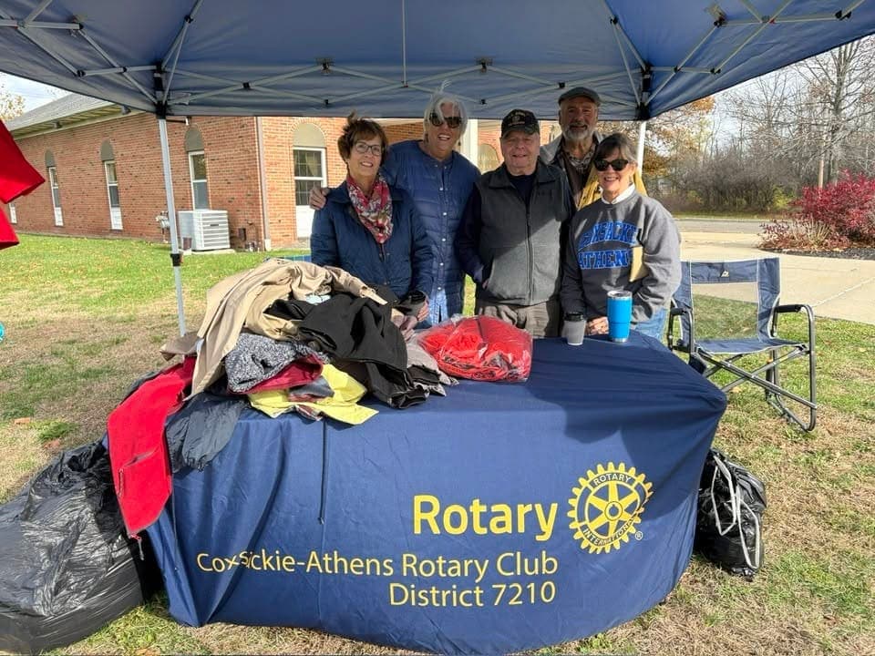 Six club members standing under a canopy behind a Rotary-draped table stacked with donated coats at the club coat drive.