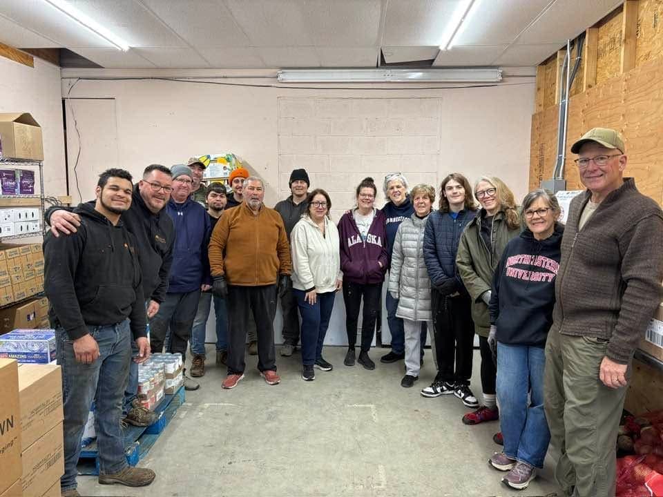 A large group of volunteers gathered among stacked crates and boxes in a local food pantry storeroom.